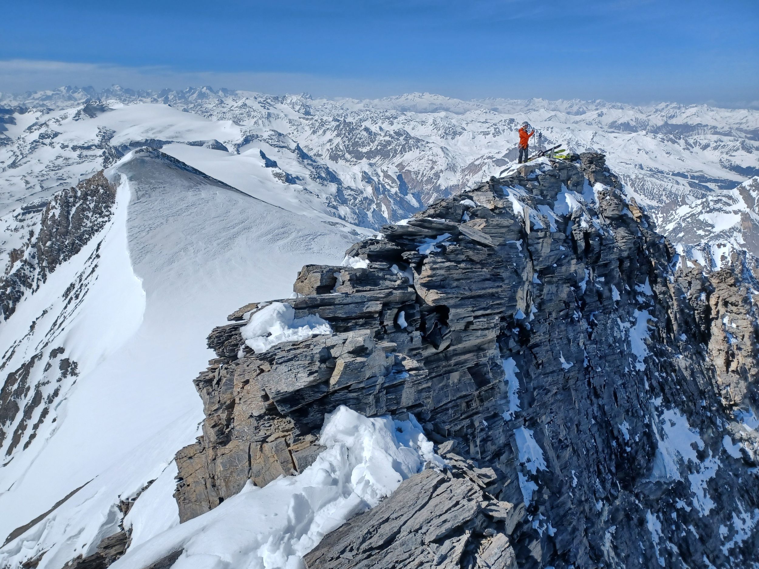 Vue sur une multitude de sommets enneigés avec un skieur sur une arête, perché au-dessus du vide.