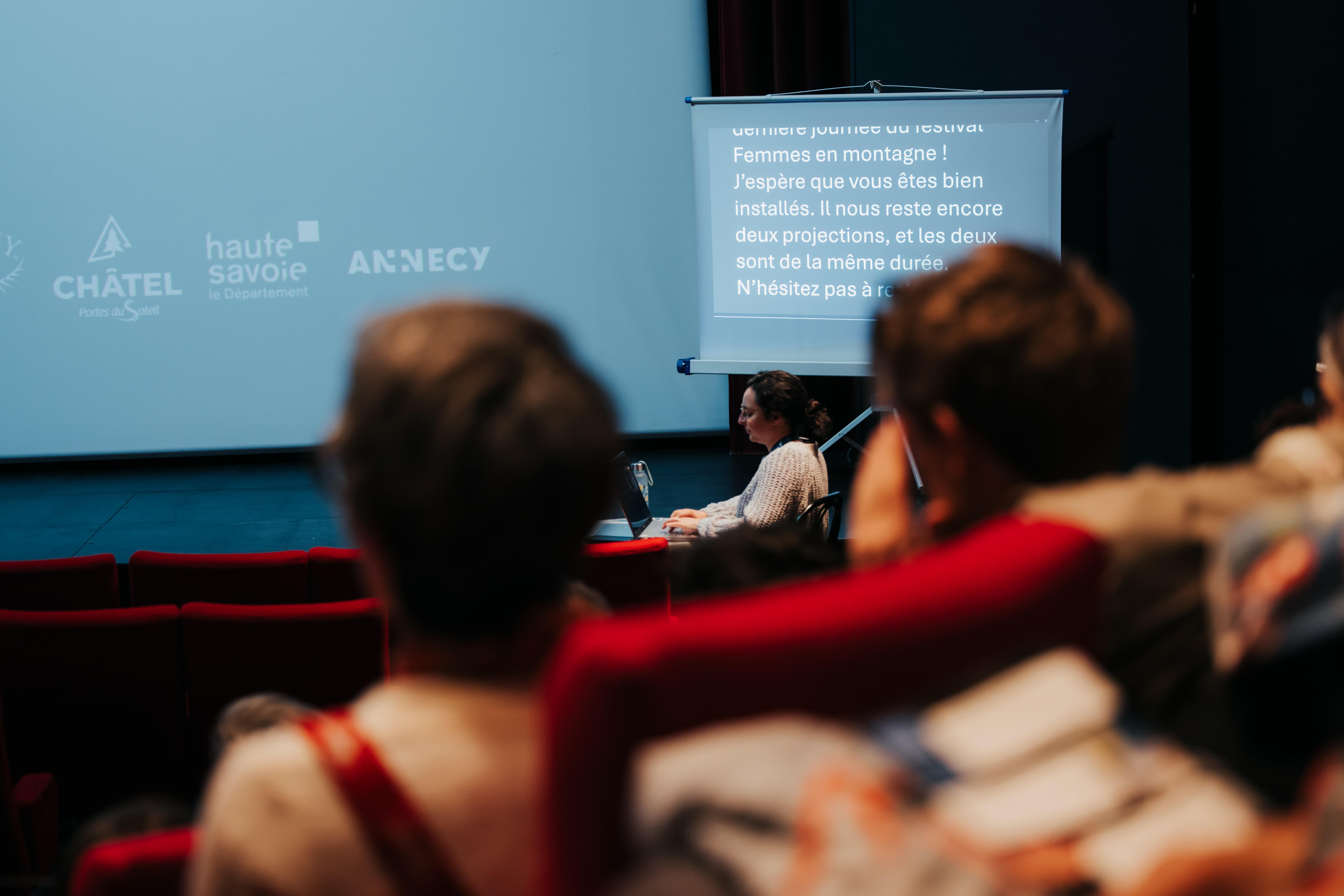 Salle de cinéma avec fauteuils rouges, public assis face à une scène. Céline est installée à une table avec un ordinateur portable pour sous-titrer l'événement. L’écran principal affiche un message d’accueil pour la dernière journée du festival “Femmes en montagne”, avec des logos de partenaires comme Châtel, Haute-Savoie et Annecy.