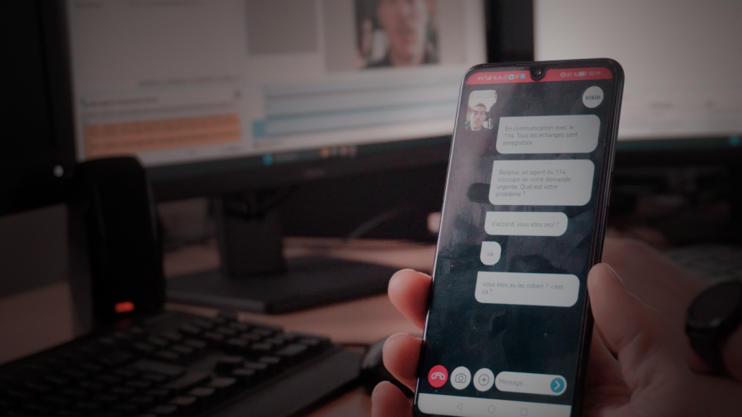 Une main tient un smartphone affichant une conversation vidéo avec un agent du 114, le fond flou montre un bureau avec un écran d'ordinateur.