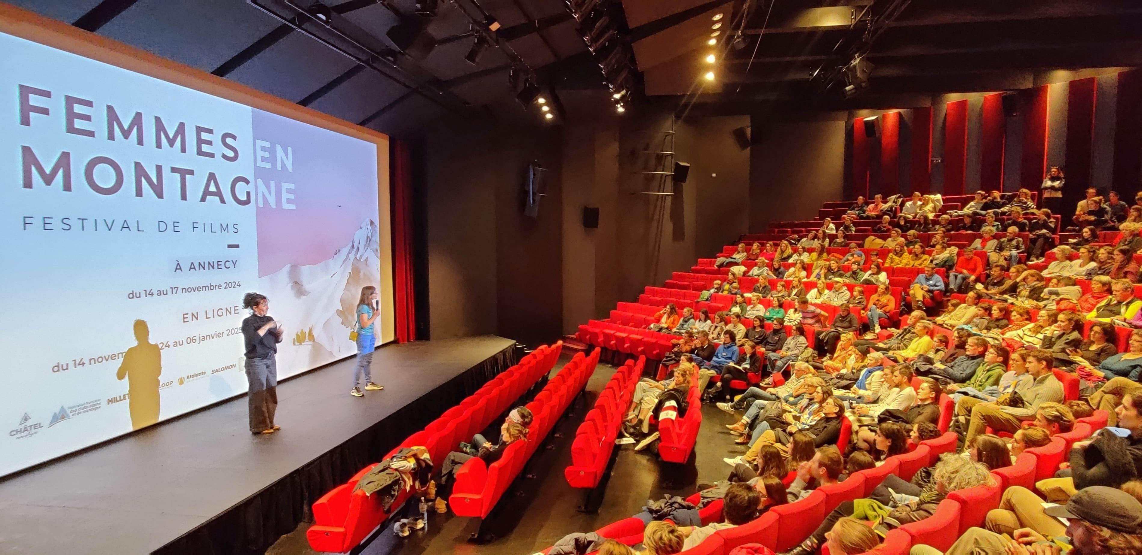Un auditorium éclairé, rempli de personnes. Le public écoute attentivement l'intervention de 2 femmes sur scène. La mention Femmes en Montagne est projeté sur l'écran de cinéma.