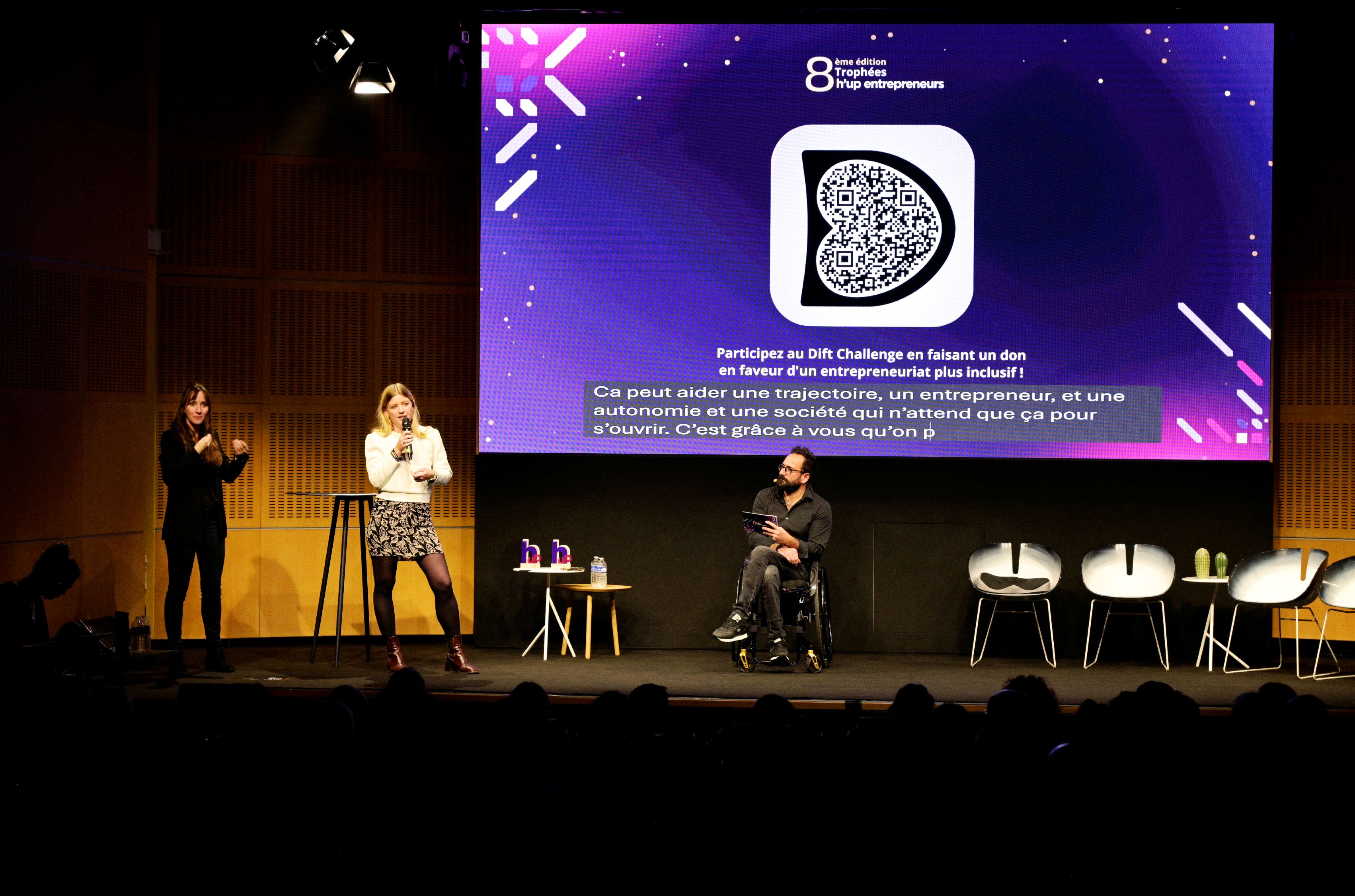 Sur scène lors des Trophées h’up entrepreneurs, une interprète en langue des signes, une personne en fauteuil roulant avec une tablette, et une intervenante au micro incarnent l’inclusion. En arrière-plan,  une présentation est projetée sur grand écran avec des sous-titres en bas de l'écran.